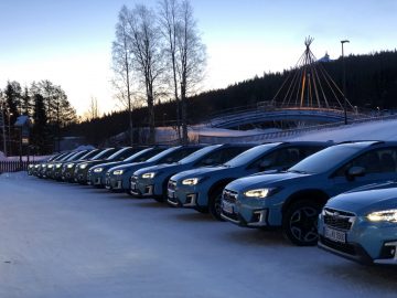 Rij auto's, waaronder een Subaru Forester, geparkeerd op een besneeuwde parkeerplaats in de schemering met bomen en een tipi-achtig bouwwerk op de achtergrond.