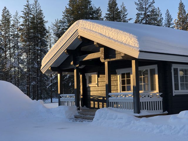 Een Subaru Forester geparkeerd voor een houten hut met een dikke laag sneeuw op het dak, omringd door een besneeuwd landschap bij zonsondergang.