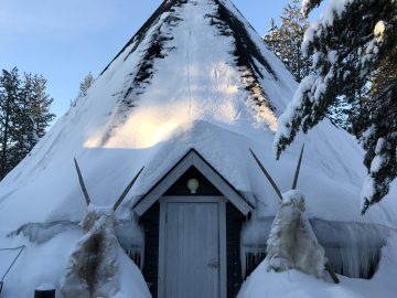 Een driehoekige houten hut bedekt met sneeuw tegen een achtergrond van bomen en een Subaru Forester onder een helderblauwe lucht.