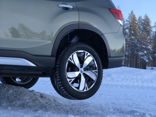Close-up van het achterwiel van een Subaru Forester met sneeuwbestendige banden op een besneeuwde weg, met bomen op de achtergrond.