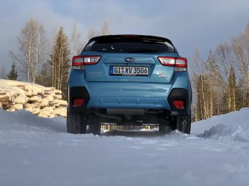 Een blauwe Subaru XV e-Boxer SUV geparkeerd op besneeuwd terrein met op de achtergrond bomen en gestapeld brandhout.