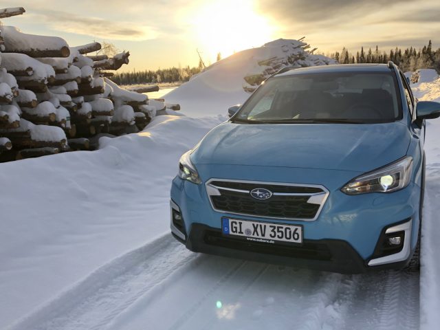 Blauwe Subaru XV e-Boxer geparkeerd op een besneeuwd pad vlakbij een stapel boomstammen met een ondergaande zon op de achtergrond.