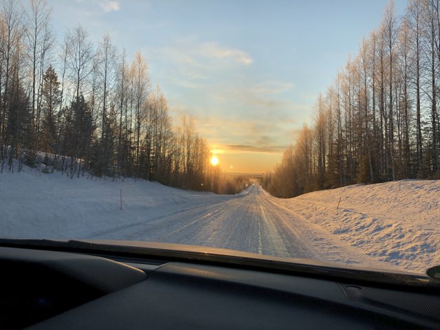 Een besneeuwde weg geflankeerd door bomen met de ondergaande zon in de verte, gezien vanaf het dashboard van een Subaru XV e-Boxer.