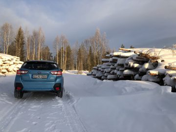 Een blauwe Subaru XV e-Boxer geparkeerd op een besneeuwde weg in de buurt van stapels gekapt hout in een bosrijke omgeving tijdens de winter.