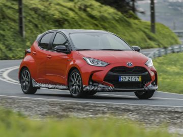 Rode Toyota Yaris hatchback auto rijden op een bochtige weg met groen op de achtergrond.