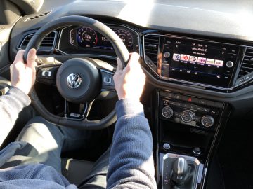 Handen van de bestuurder aan het stuur van een Volkswagen T-Roc R, met het dashboard en de middenconsole.