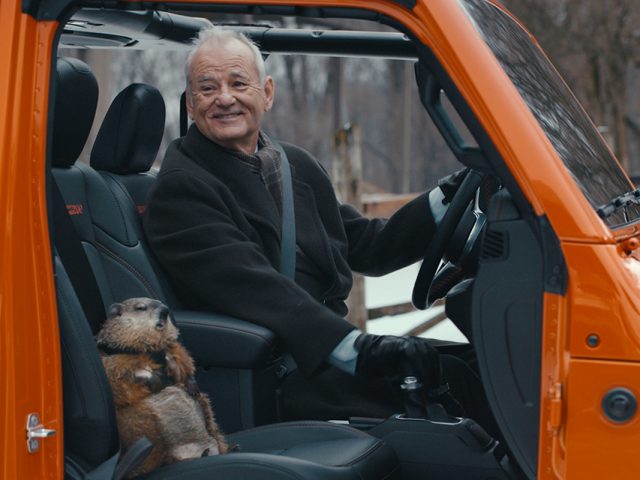 Een oudere man glimlachend naar de camera met een groundhog naast hem op de passagiersstoel van een oranje voertuig, ter ere van Super Bowl Sunday.