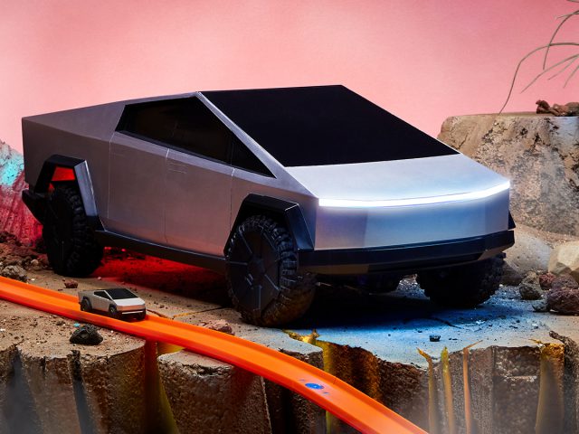 Futuristische Cybertruck op een gesimuleerd offroad-terrein met een miniatuurmodel op een speelgoedbaan op de voorgrond.