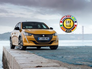 Een gouden auto geparkeerd aan de waterkant met een badge "Auto van het Jaar 2020" en een brug op de achtergrond.