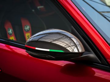 Close-up van de zijspiegel van een rode Giulia GTA met een weerspiegeling van de structuur van een gebouw.