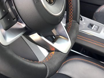 Close-up van het stuur van een auto met oranje stiksel, passend bij het stiksel van de autostoel, zichtbaar in een modern voertuiginterieur ontworpen voor snelheden tot 100 km/u.