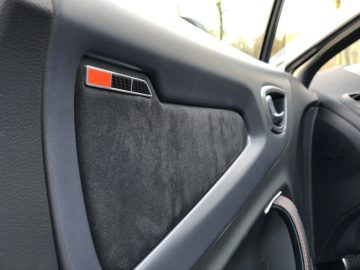 Interieur van een auto met een deel van het dashboard dat 100 km/u aangeeft en een gesloten voordeur met een zichtbare deurluidspreker en handgreep.