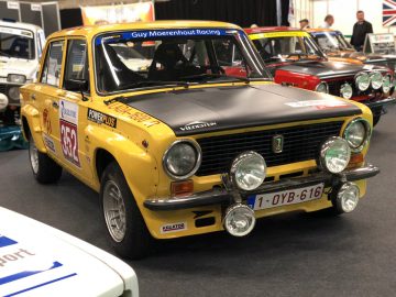 Een gele vintage rallyauto tentoongesteld op het Antwerp Classic Salon 2020, met extra koplampen en race-emblemen.