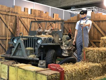 Een mannequin verkleed als piloot staat naast een vintage jeep te midden van een rustieke tentoonstelling met kippen en hooibalen op het Antwerp Classic Salon 2020.