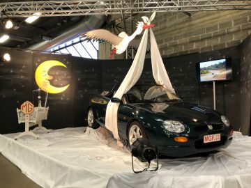 Een groene sportwagen tentoongesteld op het Antwerp Classic Salon 2020 met daarop een speels beeldhouwwerk van een vliegende ooievaar, tegen een achtergrond van een halve maan, begeleid door multimedia