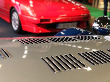 Close-up van de ventilatieopeningen aan de achterkant van een auto op het Antwerp Classic Salon 2020, met een rode sportwagen op de achtergrond.