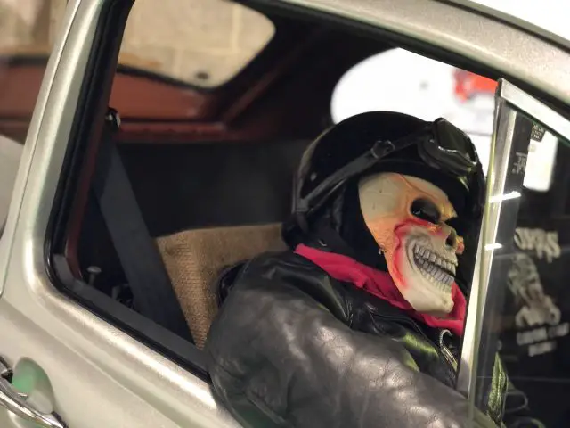 Een persoon met een schedelmasker en motorhelm zittend op de bestuurdersstoel van een auto op het Antwerp Classic Salon 2020.