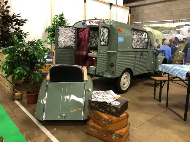 Vintage groene camper tentoongesteld op het Antwerp Classic Salon 2020 met bijpassende minitrailer, omringd door klassieke reisaccessoires zoals koffers en een plant.