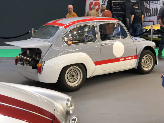 Een vintage Fiat Abarth met racestrepen tentoongesteld op het Antwerp Classic Salon 2020, met de achterste motorruimte open en mensen op de achtergrond.