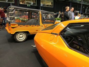 Oranje klassieke auto tentoongesteld naast een klein voertuig met een tuindisplay erin, op het Antwerp Classic Salon 2020, terwijl de aanwezigen rondlopen.