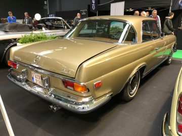 Een klassieke goudkleurige Mercedes-Benz 280SE coupé te zien op de autoshow Antwerp Classic Salon 2020, gezien vanuit de achterste driekwart hoek.
