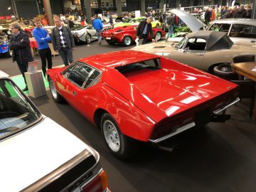 Klassieke rode sportwagen tentoongesteld op het Antwerp Classic Salon 2020, met andere vintage voertuigen en aanwezigen op de achtergrond.