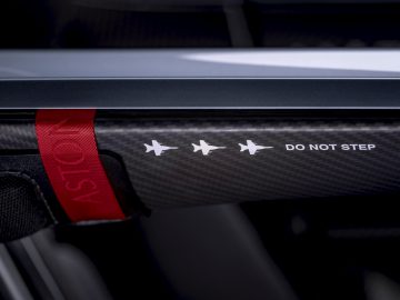 Close-up van een Aston Martin V12 Speedster koolstofvezel voertuigonderdeel met een rode trekband en een waarschuwingslabel "stap niet" naast vier witte stersymbolen.