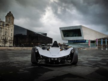 Een laaghangende BAC Mono-sportwagen geparkeerd op een stedelijk plein met donkere wolken erboven, en het museum van Liverpool op de achtergrond.