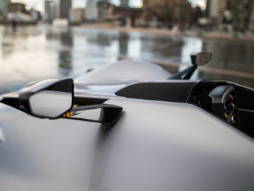 Close-up van een BAC Mono-sportwagen met focus op de zijspiegel, tegen een wazige stadsachtergrond.