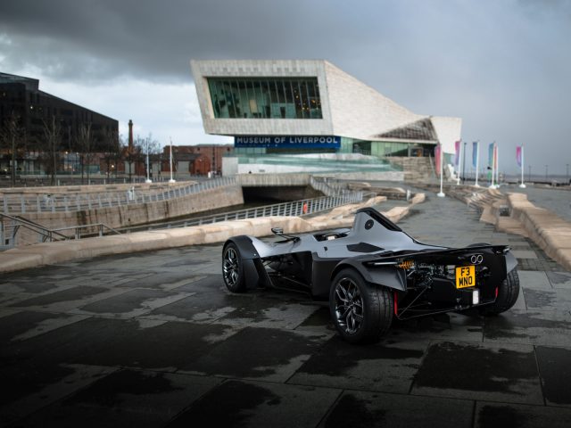 Een zwarte BAC Mono-sportwagen geparkeerd op een natte promenade met het Museum van Liverpool op de achtergrond op een bewolkte dag.