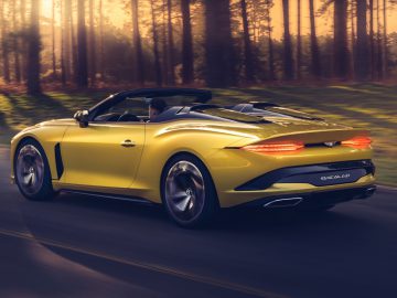 Een gele Bentley Mulliner Bacalar converteerbare sportwagen die bij zonsondergang op een weg door een bos rijdt.