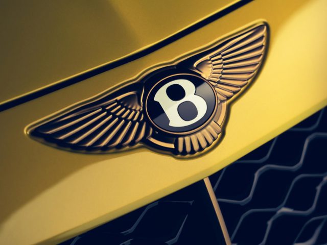 Close-up van een gouden Bentley Mulliner Bacalar-badge met vleugels en het nummer 8 op de motorkap van een gele auto.