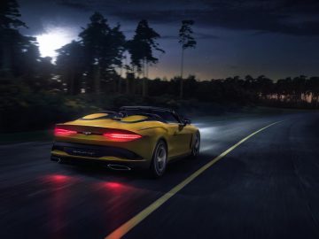 Een gele Bentley Mulliner Bacalar converteerbare sportwagen die 's nachts over een weg door een bos rijdt met brandende achterlichten.