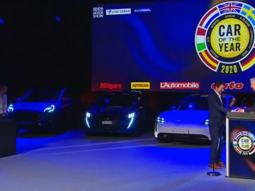 Prijsuitreiking op het evenement "Auto van het Jaar 2020" met twee personen op het podium en tentoongestelde voertuigen.