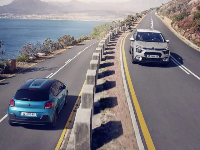Twee Citroën C3-auto's rijden langs een kustweg met bergen op de achtergrond.