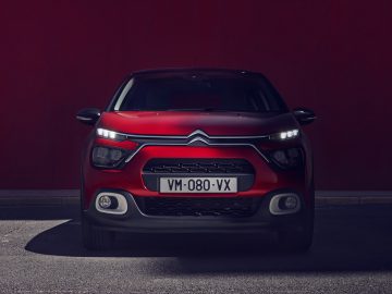 Een rode Citroën C3-auto geparkeerd voor een donkerrode muur, gedeeltelijk in de schaduw terwijl de koplampen uit zijn.