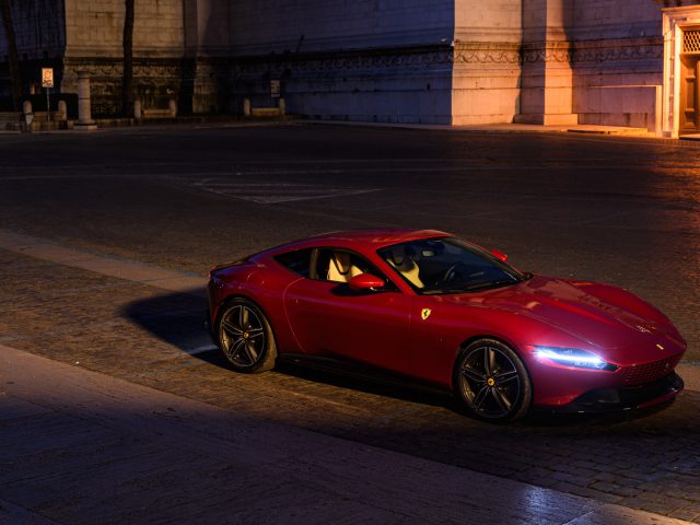 Rode Ferrari Roma geparkeerd in een stadsstraat 's nachts met gebouwen verlicht op de achtergrond.