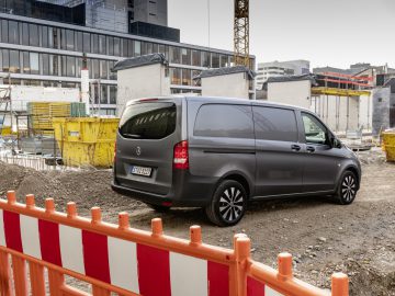 Een grijze Mercedes-Benz Vito geparkeerd vlakbij een bouwplaats met gebouwen en kranen op de achtergrond.