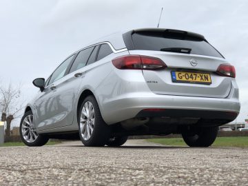 Zilverkleurige Opel Astra Sports Tourer geparkeerd op een verhard terrein met een grasachtige achtergrond.