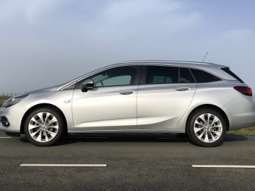 Zilverkleurige Opel Astra Sports Tourer geparkeerd aan de kant van een weg met een heldere hemel op de achtergrond.