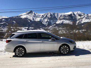 Zilveren Opel Astra Sports Tourer geparkeerd op een besneeuwde weg met bergen op de achtergrond.