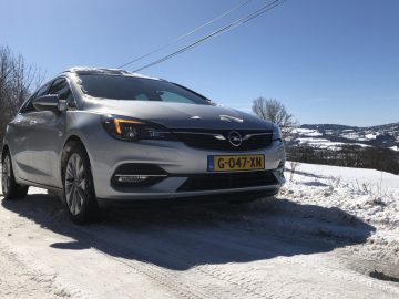 Zilverkleurige Opel Astra Sports Tourer geparkeerd op een besneeuwde weg met een helderblauwe lucht en heuvelachtig landschap op de achtergrond.