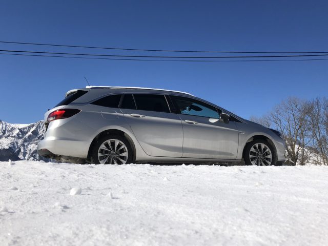 Een zilveren Opel Astra Sports Tourer geparkeerd op een met sneeuw bedekt terrein met bergen op de achtergrond onder een helderblauwe lucht.