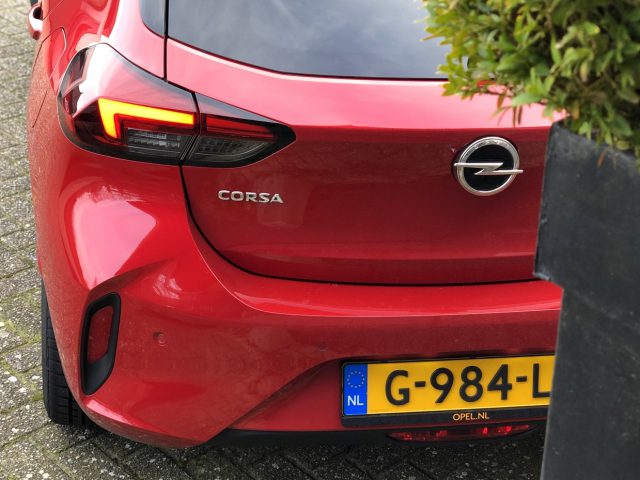 Rode Opel Corsa-auto geparkeerd op straat, gedeeltelijk zicht vanaf de rechterachterzijde.