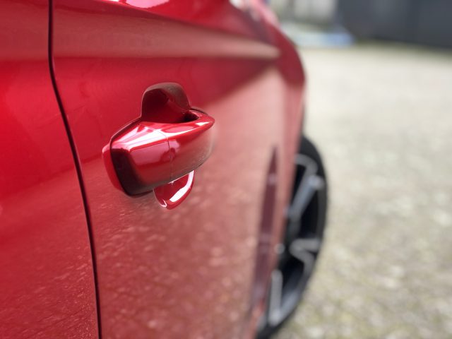 Close-up van de deurklink van een rode Opel Corsa met waterdruppels, onscherpe achtergrond.