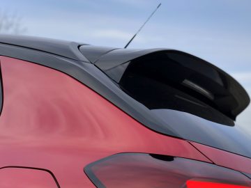 Close-up van het achterste gedeelte van een rode Opel Corsa met een spoiler en antenne tegen een bewolkte hemelachtergrond.