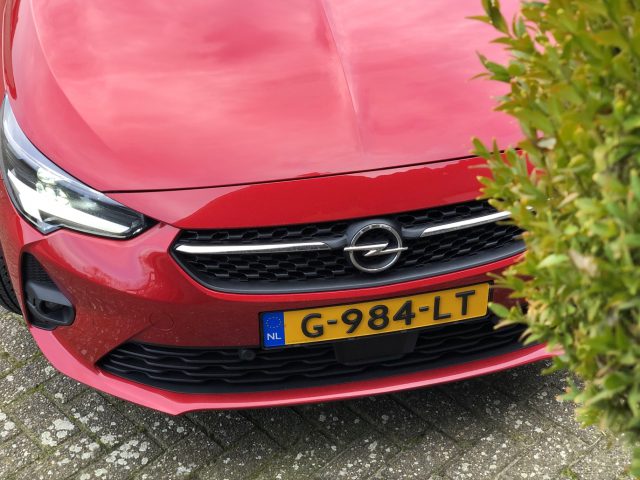 Een close-up van de grille en gedeeltelijke motorkap van een rode Opel Corsa met een Nederlands kenteken.
