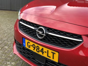 Close-up van de grille van een rode Opel Corsa met het logo van het merk en een Nederlands kenteken.