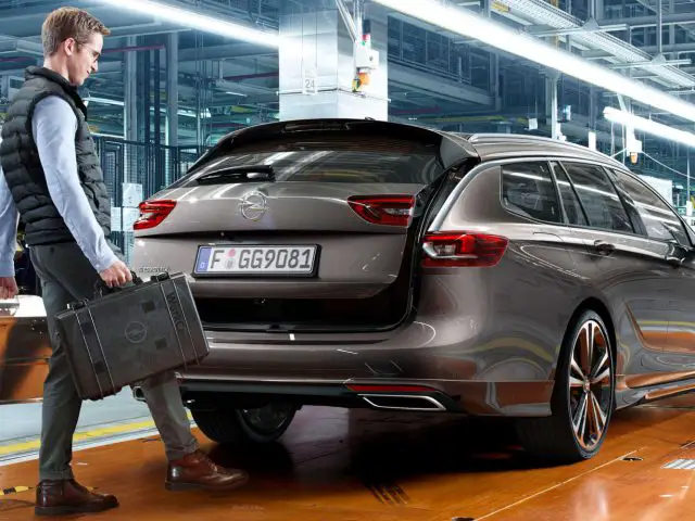 Een man met een koffertje loopt langs een geparkeerde Opel Insignia in een industriële faciliteit.