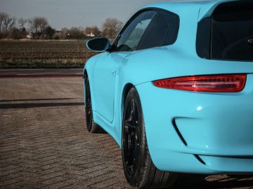 Lichtblauwe Porsche Boxster Shooting Brake geparkeerd op een verhard terrein met velden op de achtergrond.